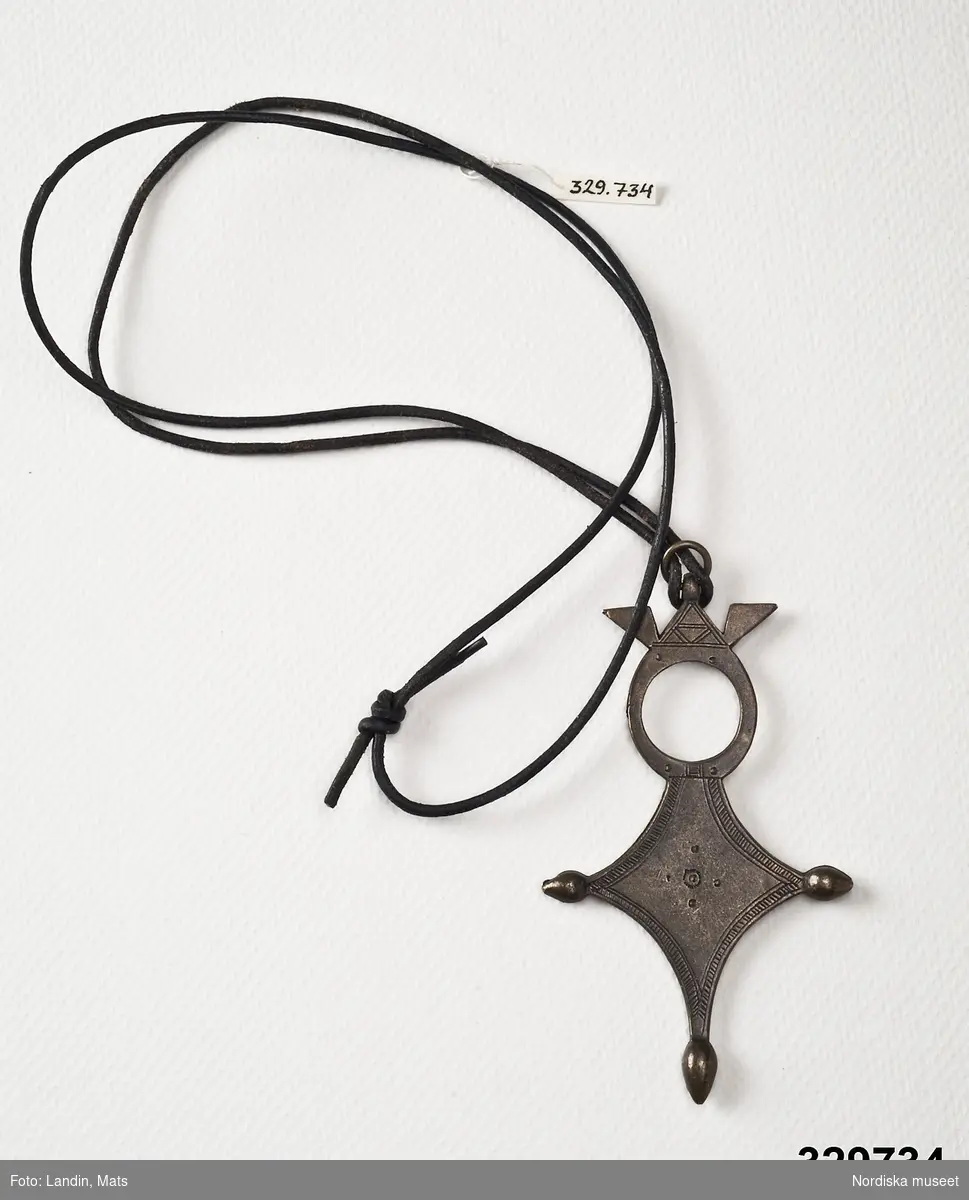 Halsband, smal svart läderrem med hänge i mörk metall korsformig med rund genombruten överdel. Stämpel med texten "Scooter Paris" på baksidan.
/Zingoalla Rosenqvist 2009-02-05