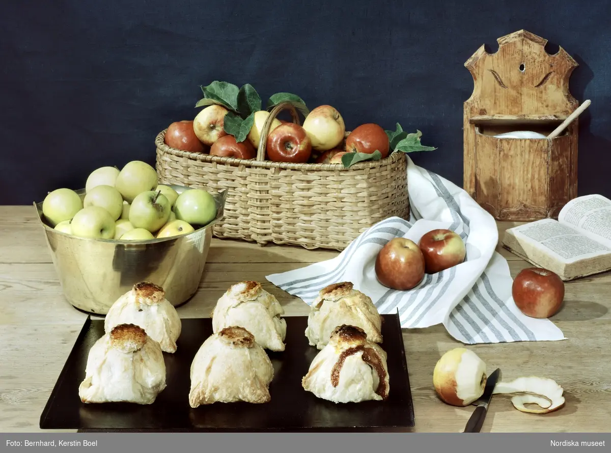 Bakning; vetebröd; inbakade äpplen. Illustration till kokbok. Äppelknyten på plåt, skål och korg fyllda med äpplen, kökshanduk, sockerburk.