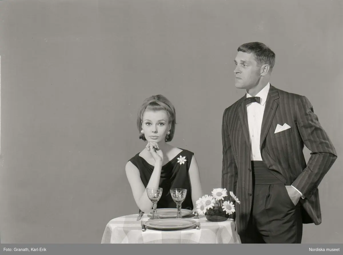 En man och en kvinna vid ett runt bord. Kvinnan sitter ned, bär en mörk överdel, brosch och örhängen. Mannen står upp, klädd i Smoking. På bordet finns en rutig duk, två tallrikar, två glas, bestick och en blomvas.