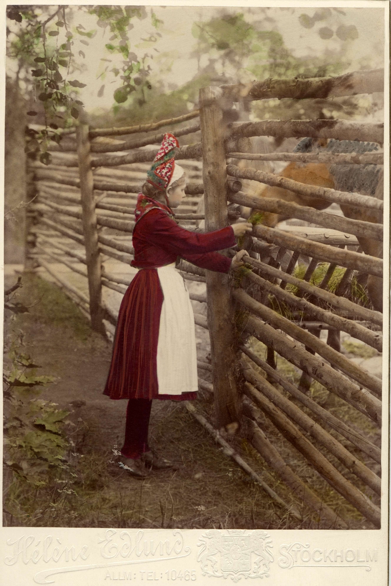En flicka iklädd folkdräkt står framför en hästhage. Handkolorerat fotografi.