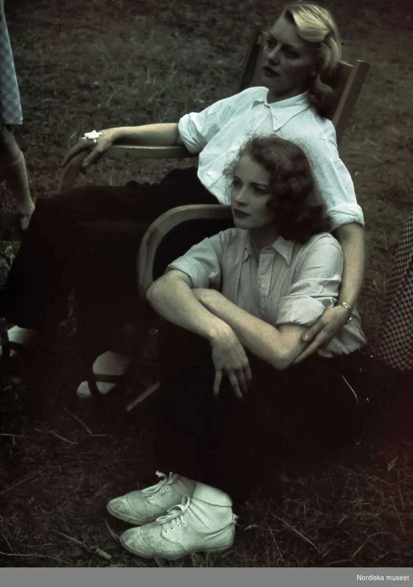 En kvinna sitter i en stol och håller om en kvinna som sitter i gräset bredvid stolen.
