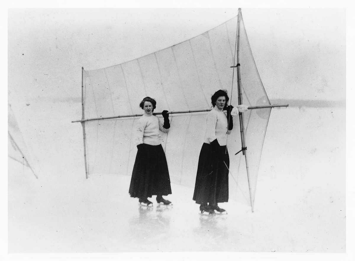 Skridskosegling på is. Två kvinnor i tidens sportkläder, långa kjolar och polotröjor, och på skridskor håller gemensamt upp ett skridskosegel bakom sig.