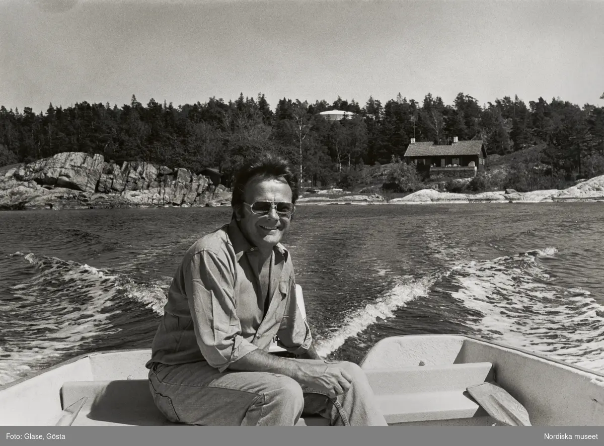 Porträtt av skådespelaren och filmregissören Gunnar Hellström.(1928-2001). Hellström sitter leende och avspänd, iklädd jeans och uppknäppt skjorta, i aktern på en liten motorbåt. I bakgrunden syns Torös klippor, ett hus vid stranden och uppe i skogen anas taket på Villa Fjolle, även kallad Villa Spies. 
