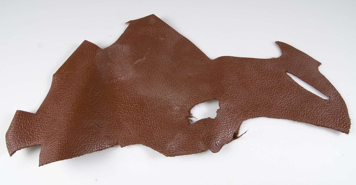 Läderbit ur vilken ovanläder till pjäxor eller dylikt skurits ut. Rödbrunt läder med strukturerad yta. Oregelbunden form.