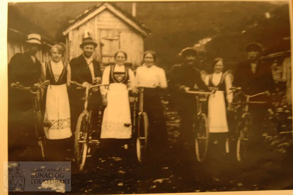 Kopi av svart/kvitt fotografi av fire karar med sykkel, tre jenter i nasjonaldrakt og ei jente i kvit bluse og mørkt utskrått skjørt. Eit hus i bakgrunn.