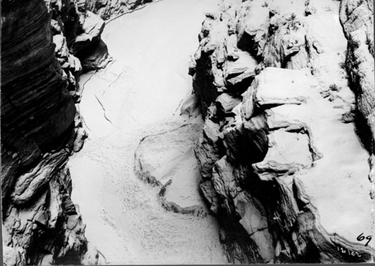 Harpefossen i Gudbrandsdalslågen, i Sør-Fron kommune i Oppland fylke.  Fotografiet er antakelig tatt fra vegbrua og ned mot det is- og snødekte elvefaret, hvor vannet inntil det ble utført reguleringsarbeidfer her i 1960-åra gikk i store og stride strømmer i sommerhalvåret.  Fotografiet viser at elveløpet gikk mellom stupbratte bergvegger.  

Før Harpefossen ble utbygd i 1960-åra var det et fall på cirka 25 meter over en strekning på om lag en kilometer her.  Fossen startet med et stup i elveleiet like ved garden Solbrå.  Nedenfor smalnet elveløpet i ei bergkløft med strid strøm og nesten loddrette vegger.  «Elveleiet i fossen er saa trangt, at vandet under flom hvirvles op langs de bratte fjeldvægge; der er store jettegryder», forteller geologen og topografen Amund Helland i sin beskrivelse av vassdraget (1913).  Det er innlysende at tømmerfløting i et slikt elveleie var både besværlig og farlig.  Helland framstilte det slik: «Laagens bredde er i Harpefoss 15-20 m.  Fossen er ofte besværlig for tømmerflødningen.  Naar der er lidet vand, bindes tømmeret let.  Under høivand gaar flødningen noksaa godt. »