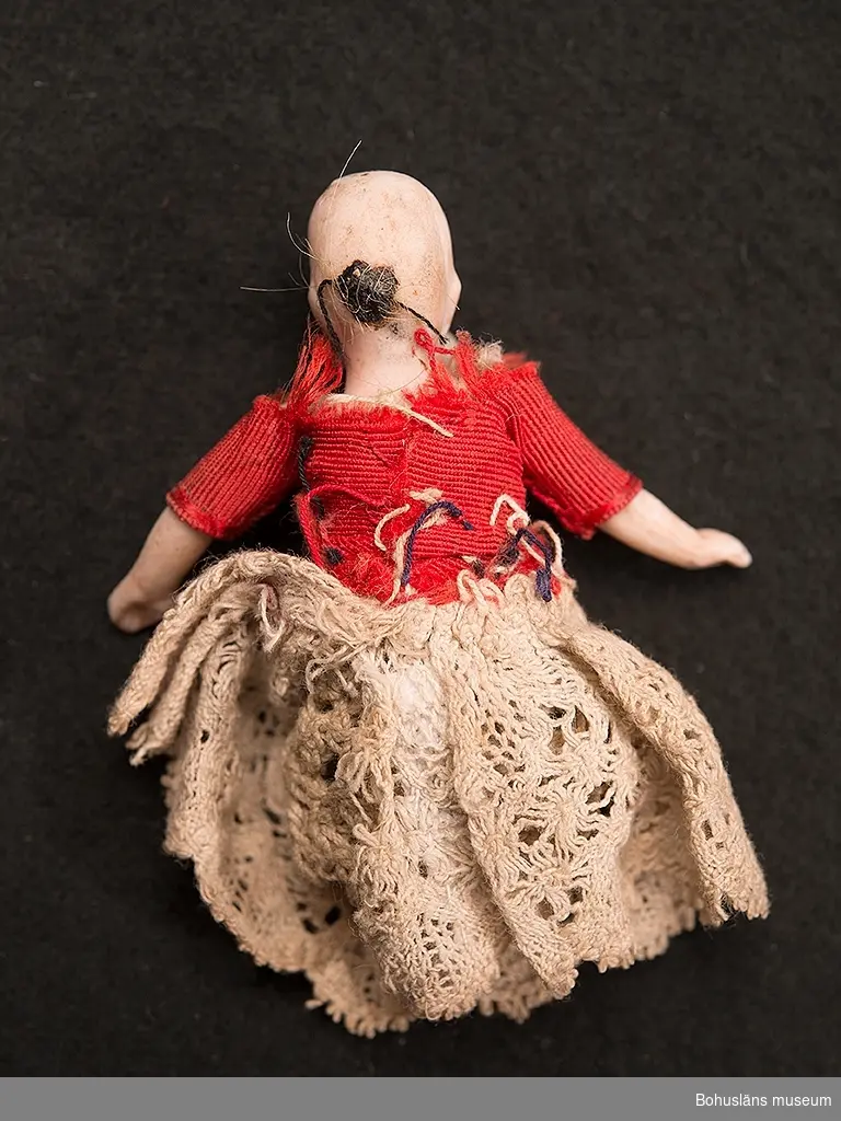 Liten docka i porslin med ledade ben och armar. Spetskjol och blus i röd ripssiden, virkad spetsunderkjol. Dockans ben är avbrutna.
Dockskåpsdocka.