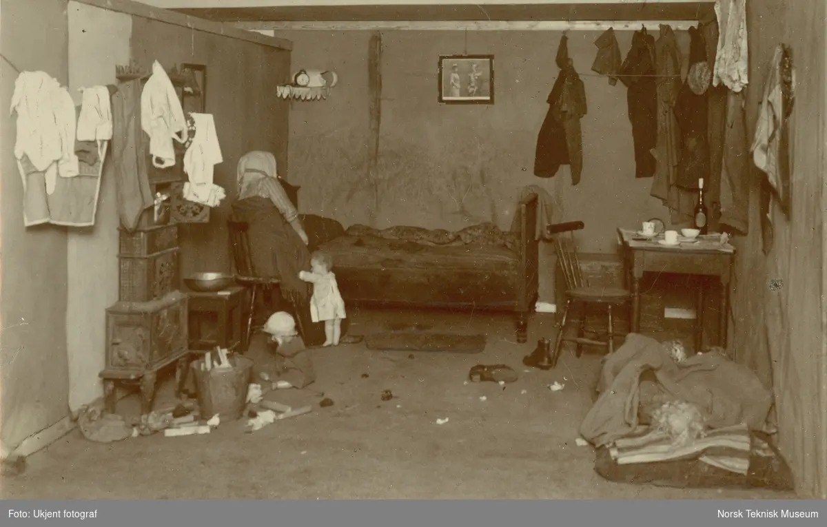Arbeiderhjem på Sagene, Oslo. Mor med to barn fotografert i kombinert stue og soverom. Bilde brukt i Barselhjemustillingen 1916