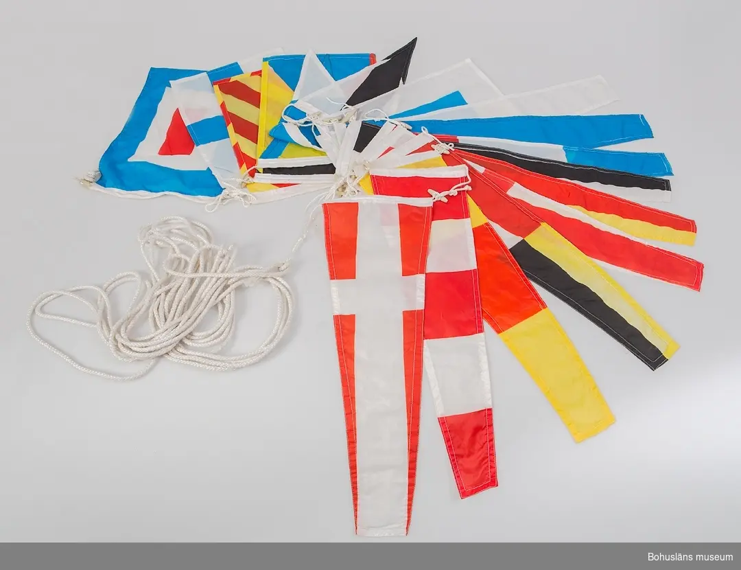 Del av internationellt signalställ på lina med 18 sydda flaggor i polyester. Flaggorna är 35x24 cm ca, rektangulära och trekantikga, inbördes kopplingsbara med knappar av plast.
Detta flaggställ använt vid festliga tillfällen för utsmyckning ombord.