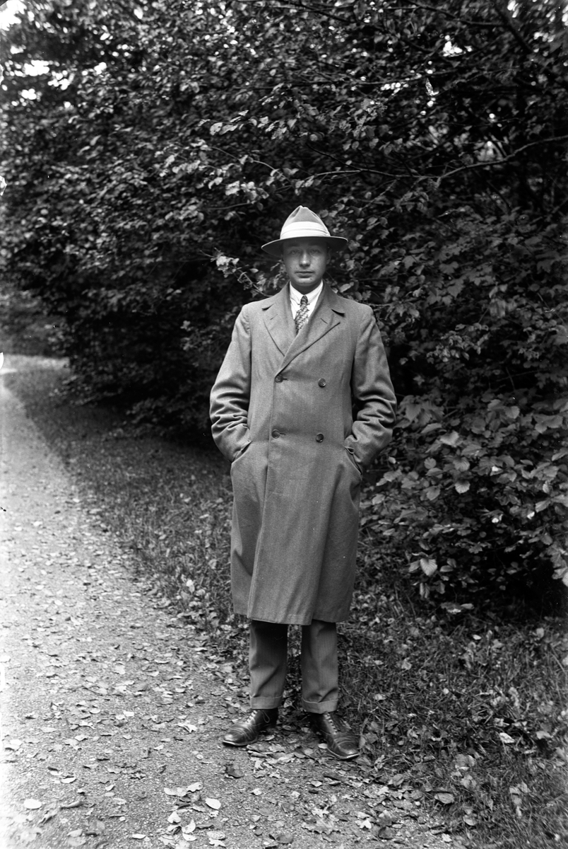 Gustav Andersson klädd i rock och hatt, står på en grusgång, vid sidan växer buskar och träd.