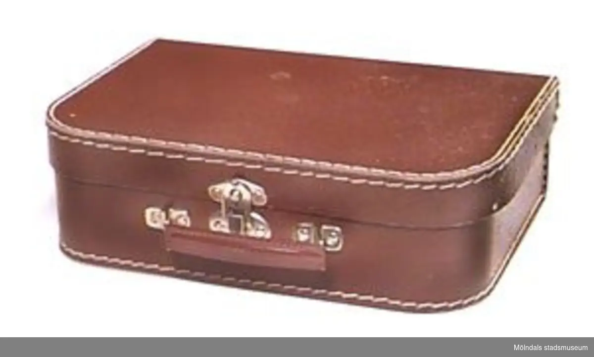 Fyrkantig brun barnresväska. Maskinsydd (med vit tråd i kanterna). Papp av unicatyp. Metallnitar och metallhandtag. Väskan är från 1950-talet. Den inköptes oanvänd av givaren till hans barn.
