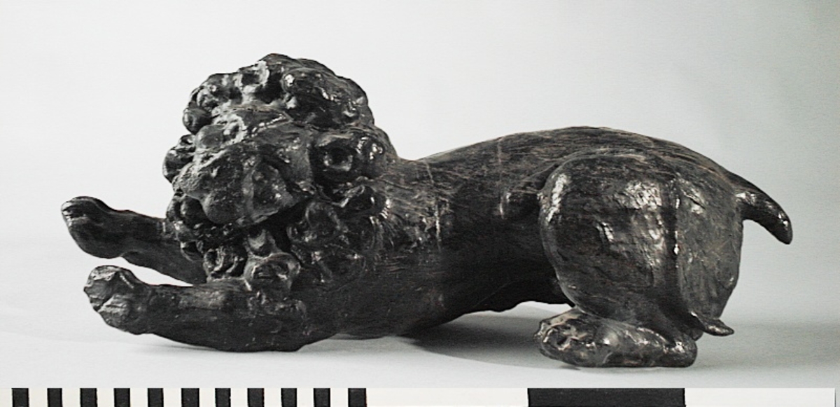 Lejon.
Skulptur av ett litet, liggande lejon med utsträckta framben och hopdragna bakben.
Lejonet är riktat mot vänster, sett från betraktaren. Det snett hållna huvudet är inramat av en yvig och krusig man och återges i det närmaste i en face. Svansen, som delvis är avbruten, är placerad över vänster lår.

Figuren är så gott som helt rundskulpterad och uppvisar ingen anliggningsyta i egentlig mening förutom mindre urtag på undersidan.
Skulpturen är relativt välbevarad. Tvärs över lejonets panna löper en spricka.

Text in English: Sculpture of a small reclining lion with its forelegs extended and hind legs tucked beneath its body.
Facing to the observer''s left, the head, held crookedly, is framed by a wavy and curly mane and is nearly en face. The tail, which has partly broken off, is held over the left hind leg.

The figure is almost three-dimensional and thus display no attachment surface except for minor grooves on the underside and the back.
The sculpture is quite well preserved.