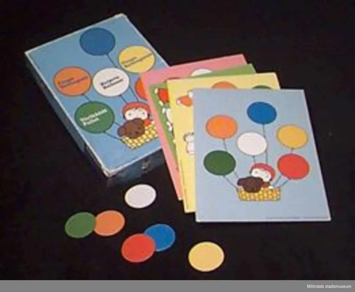 Ask innehållande spel för barn 3-7 år. Spelets namn: "Fånga ballongerna".Asken innehåller fyra spelbrickor (L: 270 mm. B: 190 mm.) i skilda färger med plats att placera ut sex ballonger på vardera bricka. Dessutom 24 ballonger (4 gula, 4 gröna, 4 vita, 4 blåa, 4 oranga och 4 röda) i form av runda pappbrickor (40 mm i diam).Spelregler och tärning saknas.Tidigare sakord: spel, leksaks-.Katrinebergs daghem var daghem.