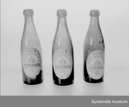 Tre flaskor tillverkade på Sundsvalls bryggeri aktiebolag. Flaskorna är bruntonade och har etiketter där texten lyder 'KLASS 2, SUNDSVALLS BRYGGERI AKTIEBOLAG, PILSNERDRICKA, SUNDSVALLS PILSNERDRICKA, SUNDSVALL'. Etiketterna har blå och röd text och motiv på vit botten och de är tillverkade hos Lithografiska AB i Norrköping.