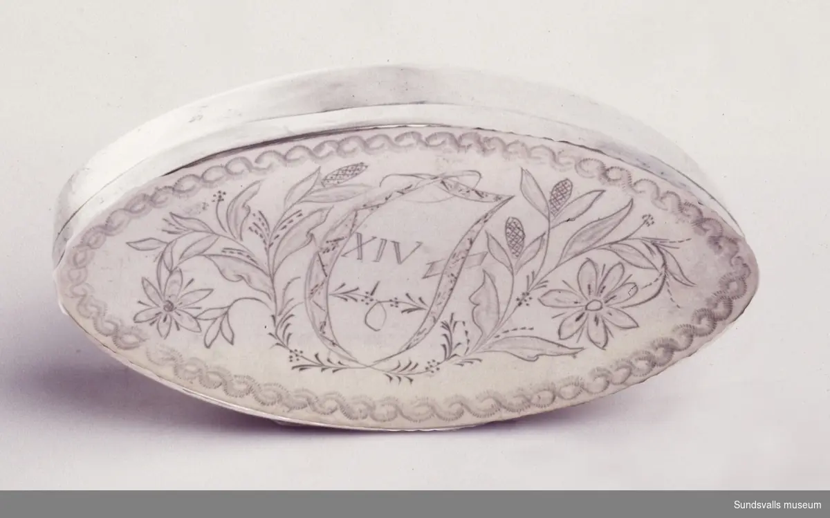 Snusdosa i silver. Spetsoval. På locket en svisselerad bård, graverade blommor, samt kungligt monogram 'C XIV J'.