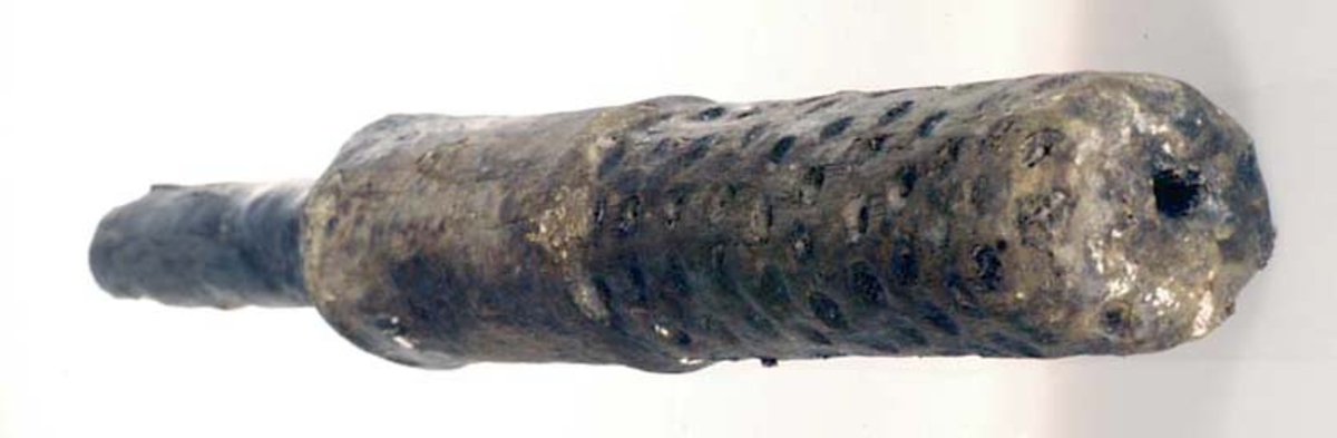 Kolv till borste. En del av ett avbrutet skaft sitter kvar i kolven. Cylindriskt formad kolv. Kolvens nedre hälft är täckt av borrade hål, en del med kvarsittande borst i sig. Den övre änden, mot skaftet till, har en större diameter. Två stycken träpluggar löper genom den övre delen för att hålla skaftet på plats.