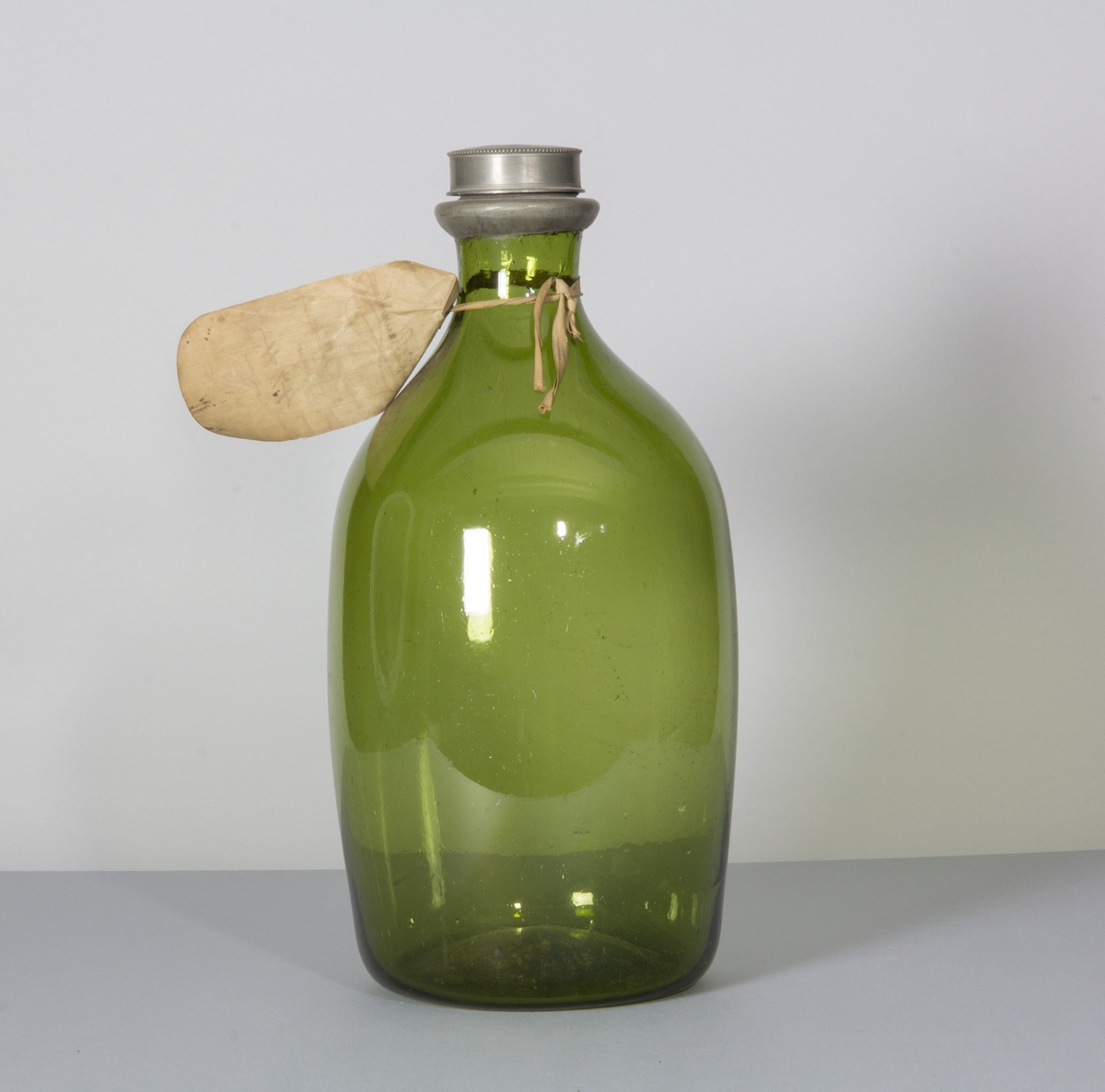 Flaska i glas, ljust grön till färgen. Rundade sidor, bukig, med låg bred hals. Skruvlock i tenn.