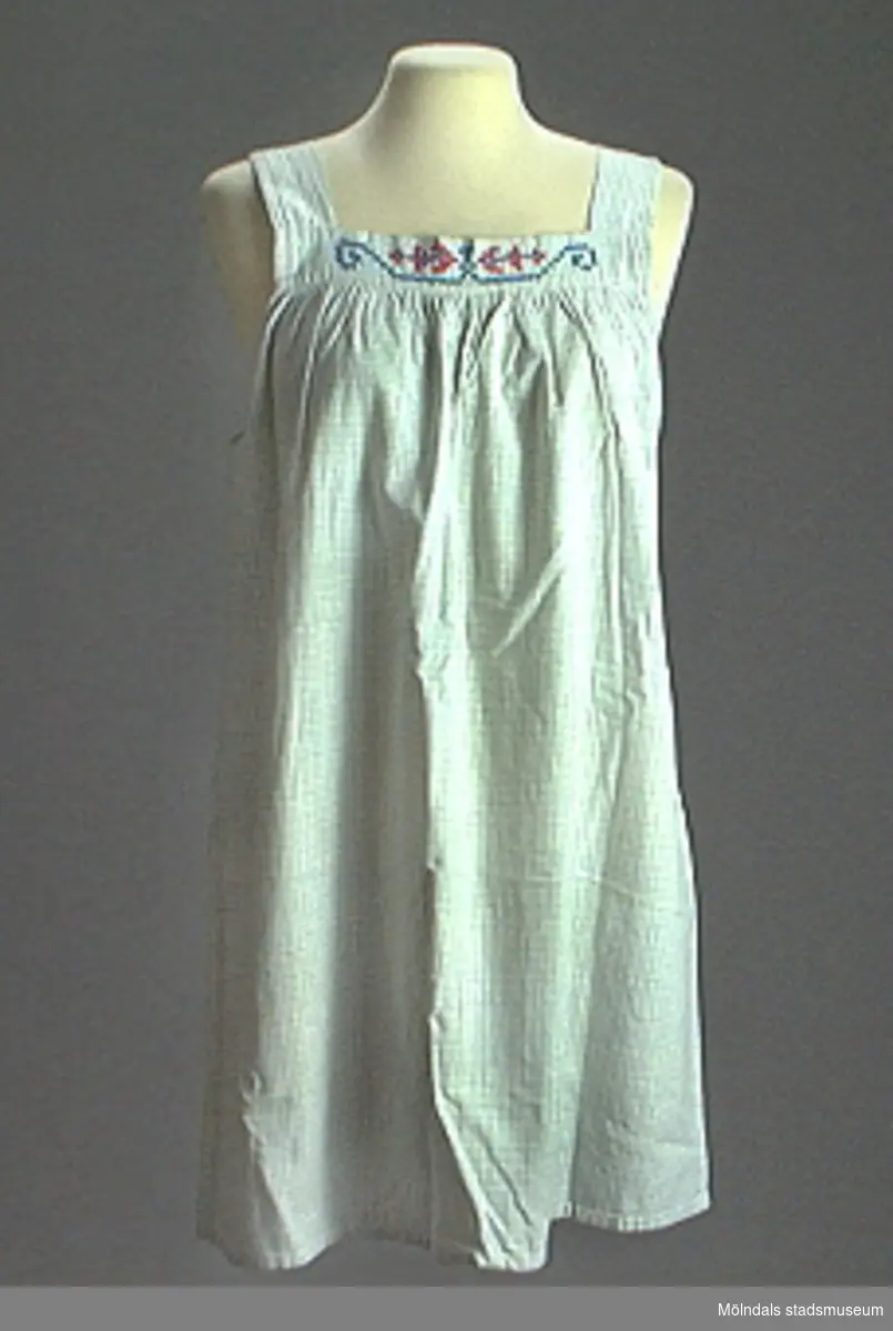 Blårutigt förkläde (21948:1), sytt som klänning med breda axelband och skärp. Broderat motiv i korsstygn mitt på livet.Förklädet något gulnat framtill. Tillhörande skärp med knapp och knapphål (21948:2).