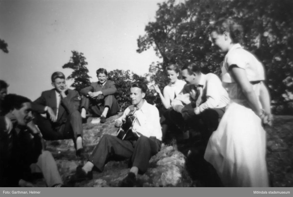 Flertalet arbetskamrater från AB Pumpindustri (API) sitter/står på en bergskulle och lyssnar till en man som spelar gitarr i mitten. Helmer Garthman och jobbarkompisar är på utflykt under 1950-talet.