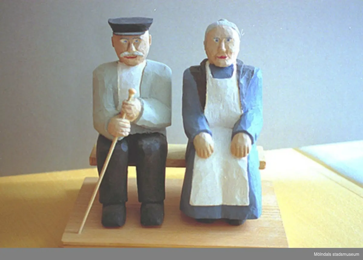 En äldre gubbe och gumma sitter bredvid varandra. Gubben har hatt och håller en käpp i handen. Gumman är klädd i vitt förkläde och har lagt sina händer i knät. Harry Bergmans "gubbar" (träfigurer).