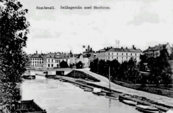 Från vänster Storbron, kv Lyckan 1, kv Penningen 1/Riksbank. I förgrunden Selångersån till vänster vedpråm och till höger båtar. Text till bild " Sundsvall. Selångersån med Storbron."