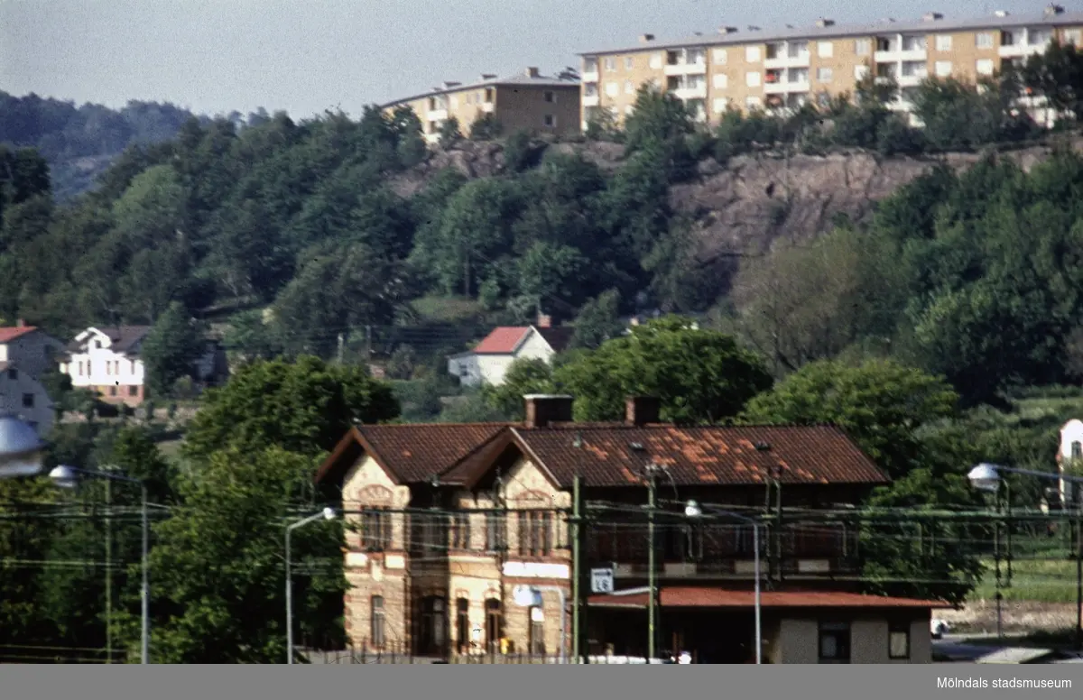 I bildens nederkant ses järnvägsstationen Mölndals Nedre vid Järnvägsgatan 1 i Mölndal, 1970-tal. I ovankant fastigheter vid Lövgatan och Delbancogatan.
