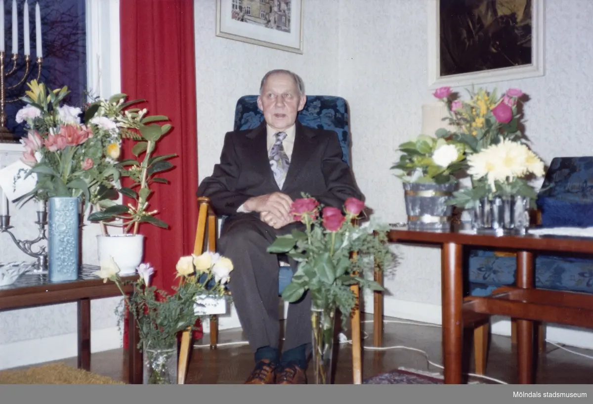 Åke Börjesson firar sin födelsedag, han är fotograferad där han sitter omgiven av blomsterbuketter i vardagsrummet. 
1970-tal.