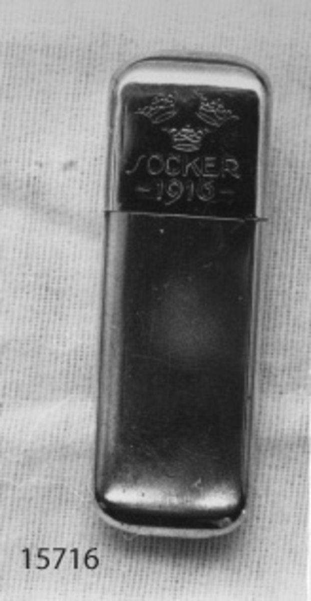 Sockerdosa av nysilver. Rektangulär. På framsidan är tre kronor instämplade, under kronorna SOCKER samt under detta -1916-. På baksidan stämplat: G.A.B. N.S.