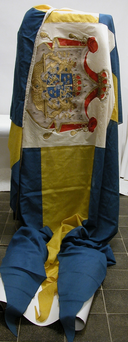 Svensk kunglig örlogsflagga i gult och blått ylletyg. Tre tungor. I det gula korsets mitt stora riksvapnet, handmålat. Liket av vitt linne, stämplad med kattfot, KUNGL. FL. STORA VAPN 5D.
