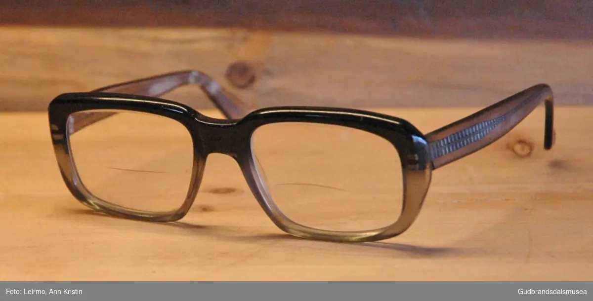Et par briller med store glass og kraftigere innfatning i hardplast, metallet i brillestengene ses gjennom siden plasten er transparent. Brillen er svart øverst og lys grågrønn/oransje rundt på innfatningen ellers. Glass med nedre lesefelt. Brukt i reparasjonsarbeid, nærarbeid.