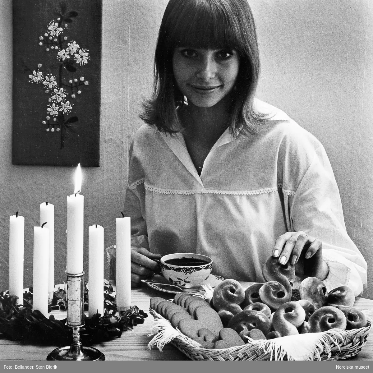 En kvinna i lucianattlinne, vid ett bord med en luciakrona, ljusstake,  korg med lussekatter och pepparkakor, samt en kaffekopp.