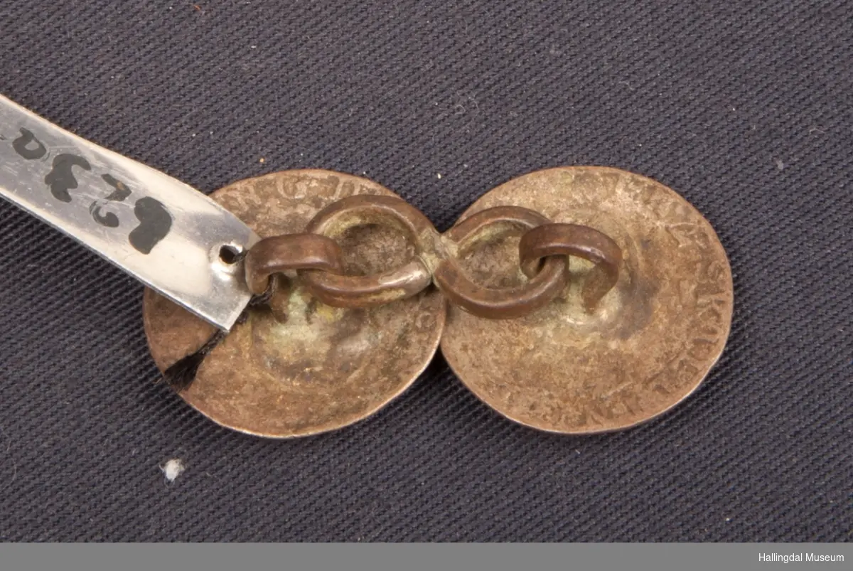 2 mynter, 2-skillingsmynter fra 1782 og 1784.  Forbundet med kringleformet sølvhempe som holder dem sammen i hempene.