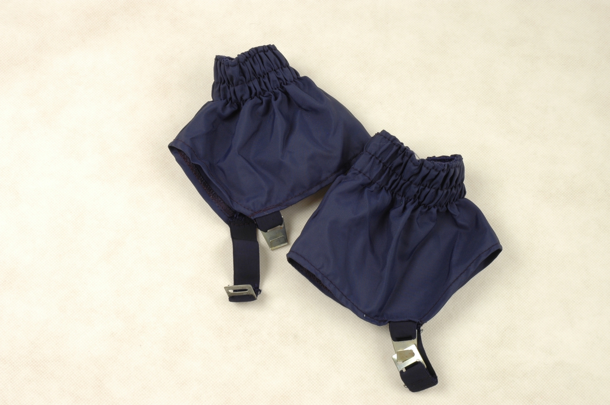 Ett par ankelhøye skigamasjer i mørkeblå nylon.  Gamasjene lukkes under skoen med strikk og hempe.