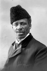 Studioportrett i halvfigur av mørkhudet mann med pelshatt. D