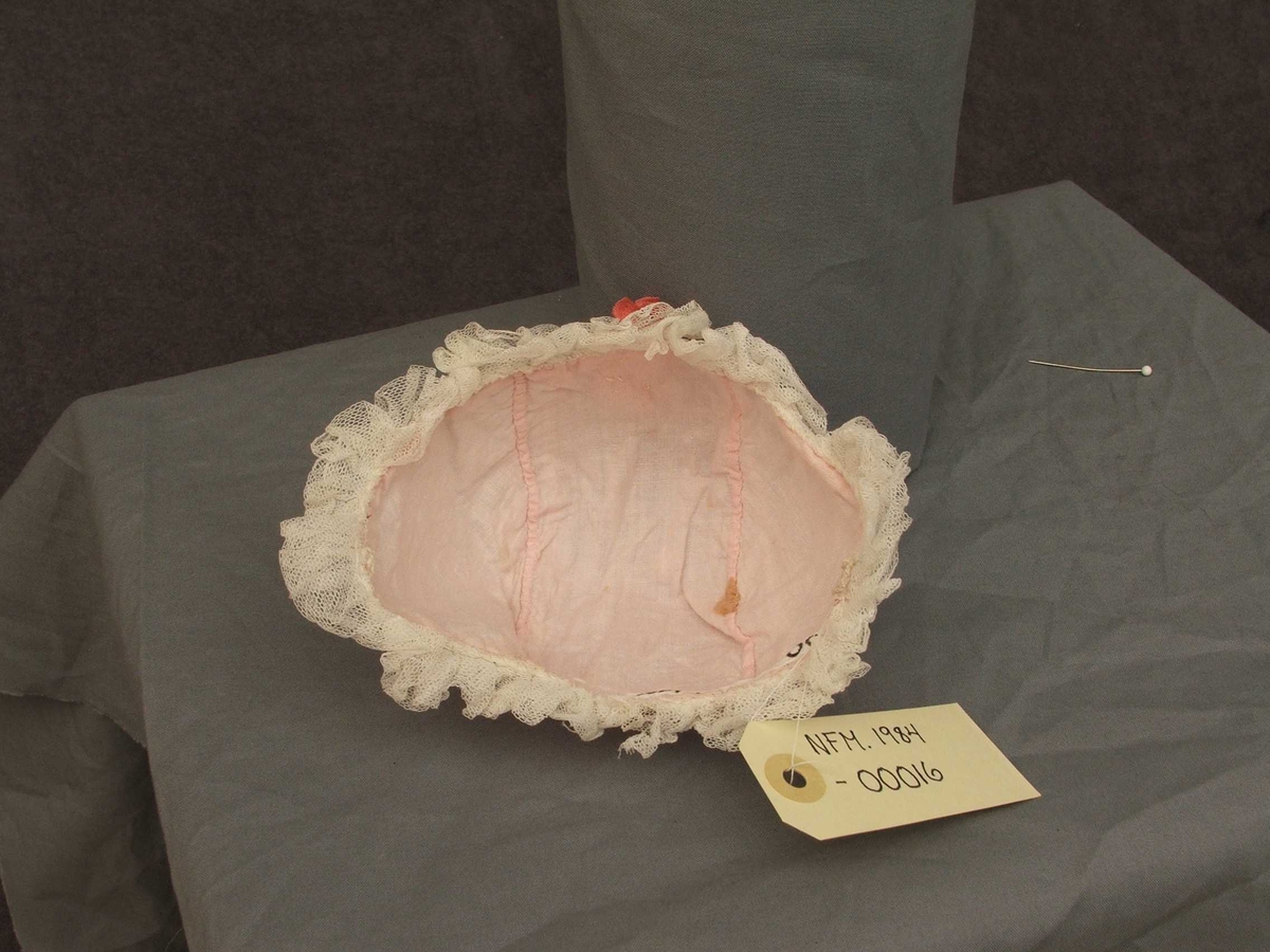 Dåpshue med tyll ytst og rosa stoff inst. Sydd saman av tre delar. Lakseraud rosett øvst på hovudet og tyllblonder rundt hovudet. Hekla hemper på kvar side.