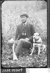 Portrett av mann med kikkert, gevær og en hund.