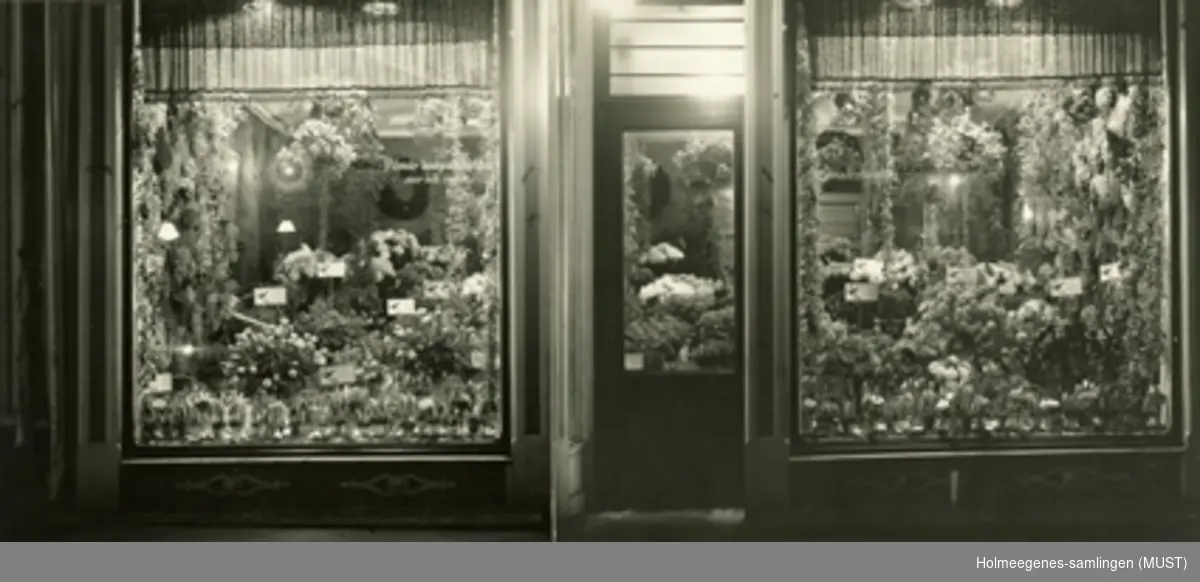 Eksteriørfotografi av en blomsterbutikk, med inngangsdør og to store utstillingsvinduer. Holmeegenes gartneris blomsterutsalg i Kirkegaten 17 i Stavanger sentrum, kalt "Blomsterhallen". Fotografi montert på kartong i glass og ramme.