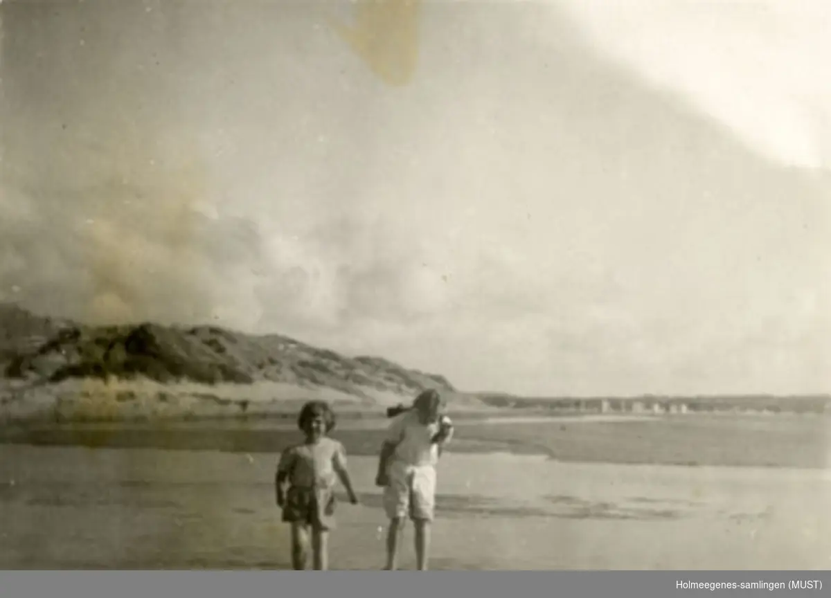 To små jenter vasser på en strand med sanddyner eller svaberg i bakgrunnen. Fint vær med lette skyer på himmelen. Antatt samme sted  og tidspunkt som ST.K.HE 2007-011-0045.