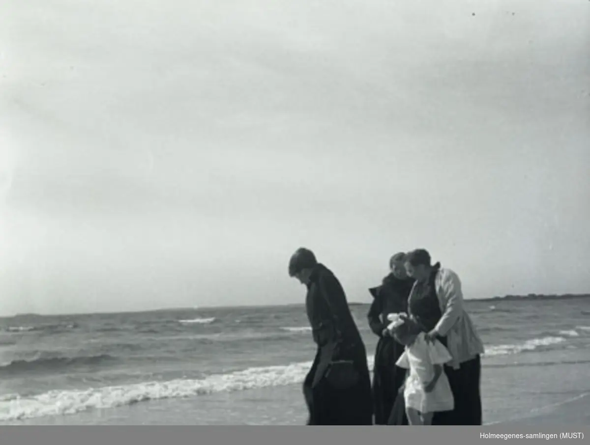 Tre kvinner og et barn på en sandstrand, utsikt mot havet. Kvinnene er kledt i mørkt yttertøy, barnet i lyst sommertøy. Muligens samme sted og tidspunkt som ST.K.HE 2007-011-0032 og -0033.