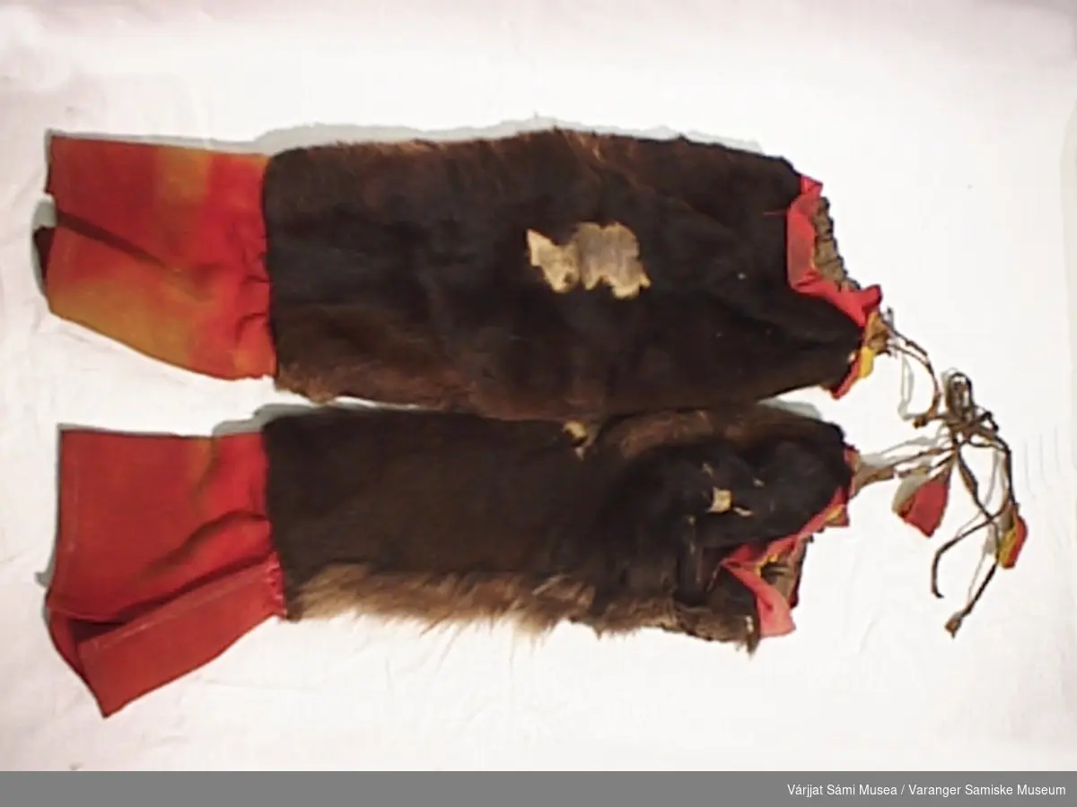 Et par herrebellinger sydd av mykt mørkebrunt skinn med pelsen på, pyntet øverst med rødt og gult klede samt en skinnanor med klededusk til stramming, rødt stykke av en annen type tøy nederst.