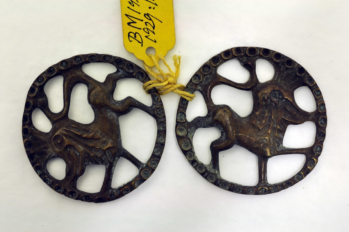 Fra protokoll: 1 par beslag av bronse (legeringsmetal). Cirkellaga sylgjebeslag i gjenombroti støypearbeid. Motiv: heraldisk løve innskrivi i cirkelring. Tvermaal 4,25 cm.