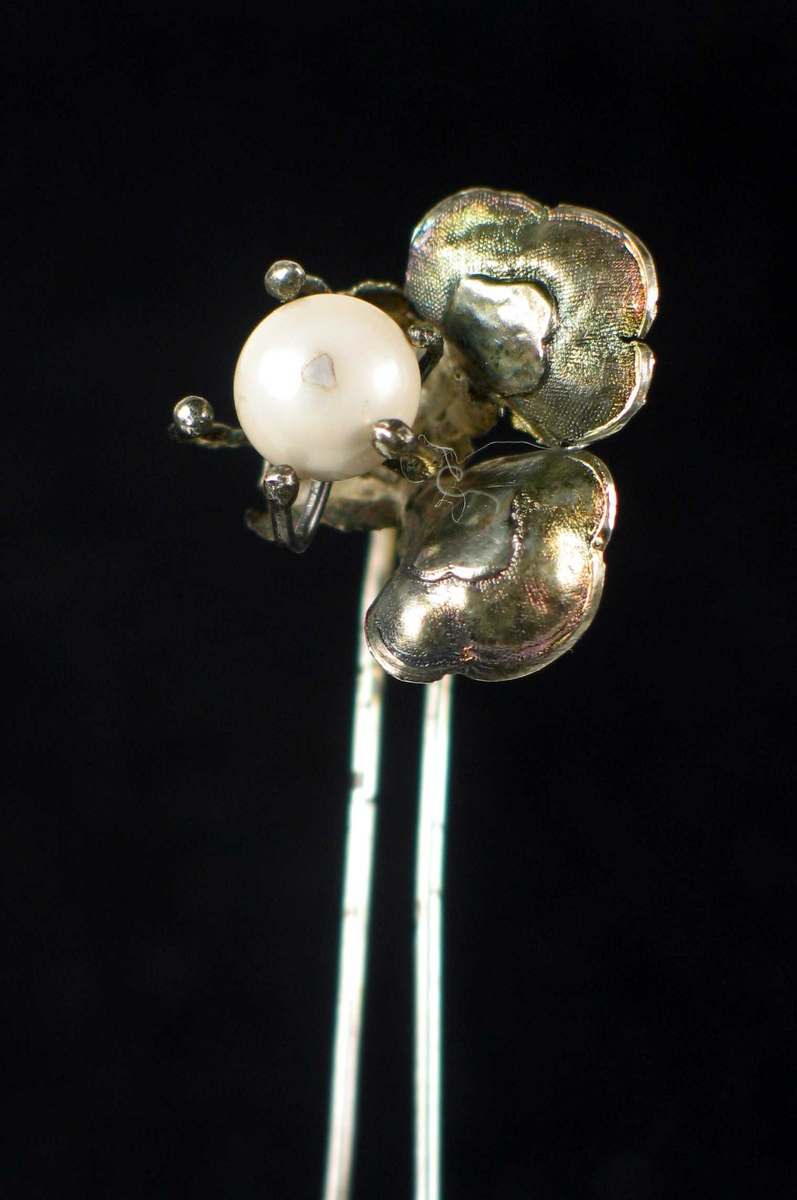 Hårpynt i sølv med tre blad og innfattet hvit perle i midten. Påloddet lang hårnål. Det ene bladet er brukket.