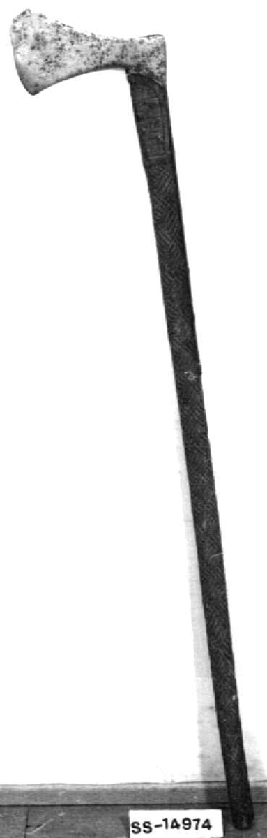Asymmetrisk økseblad, sammensveiset med overliggende jernskinne.
Fremre øksebladspiss brukket.
Rikt dekorert skaft med flettet rutemotiv. Innskripsjon på øvre del av skaftet (se Påført tekst)
