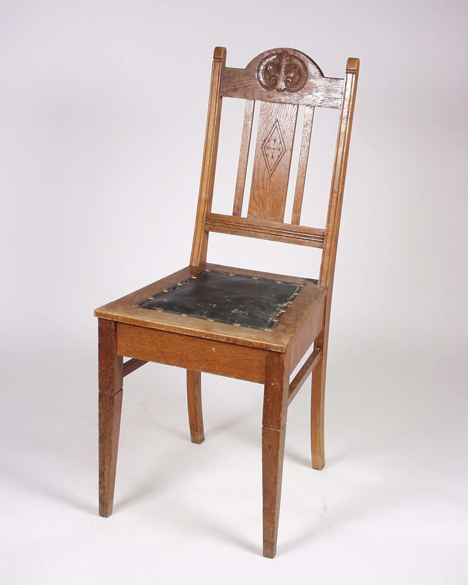 Ganske enkel stol av eik. Gjennombrutt rygg, med utskåret bladmotiv på toppstykke og ryggbrett. Sete med svart skai festet med messingbolter.