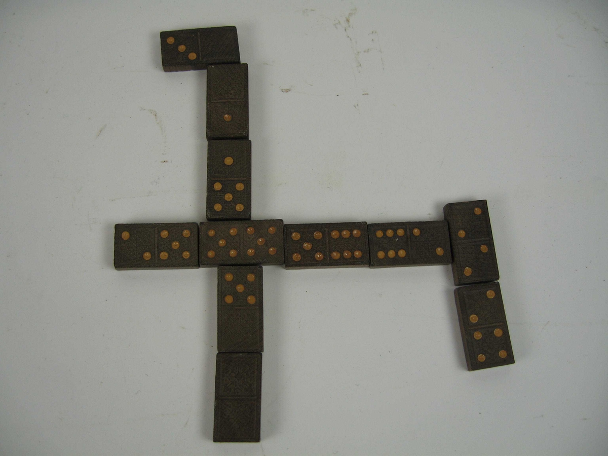 Et sett med 28 dominobrikker laget av en mørk tresort. Baksiden har et jugendmønster, mens på oversiden er nummereringen merket med oker fargeflekker.