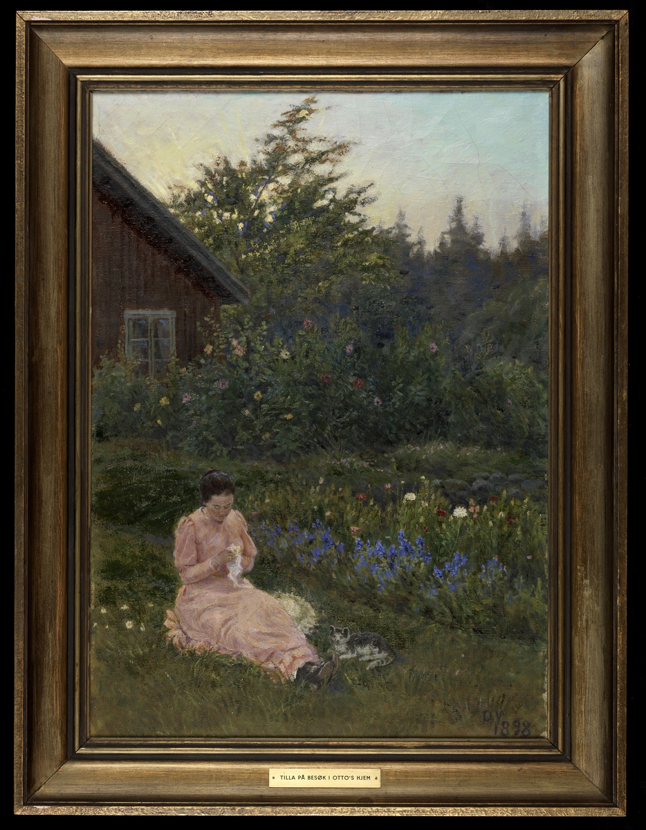 Hage, i forgrunnen Tilla Valstad i rosa kjole sittende i gresset, katt ved siden, blomsterbed, i bakgrunnen hj. av rødt hus, trær. 