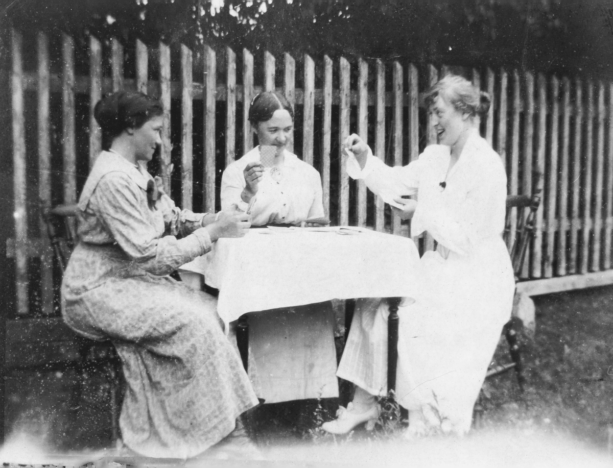 Gruppebilde,Tre damer i kjoler  spelar kort ved eit bord
Bak er ettstakittgjerde.