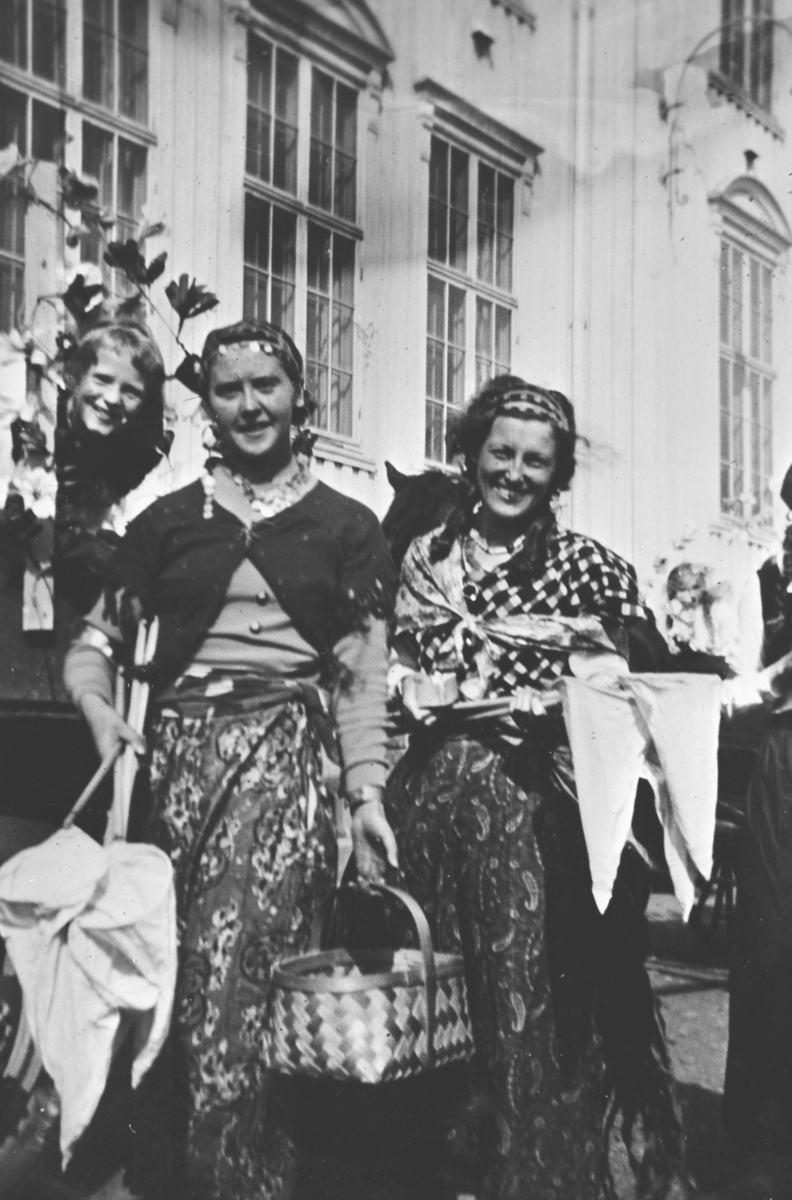 Damer i kostymer med håver og kurv foran hus. Margit Sundelin i midten. De to andre er ukjent.