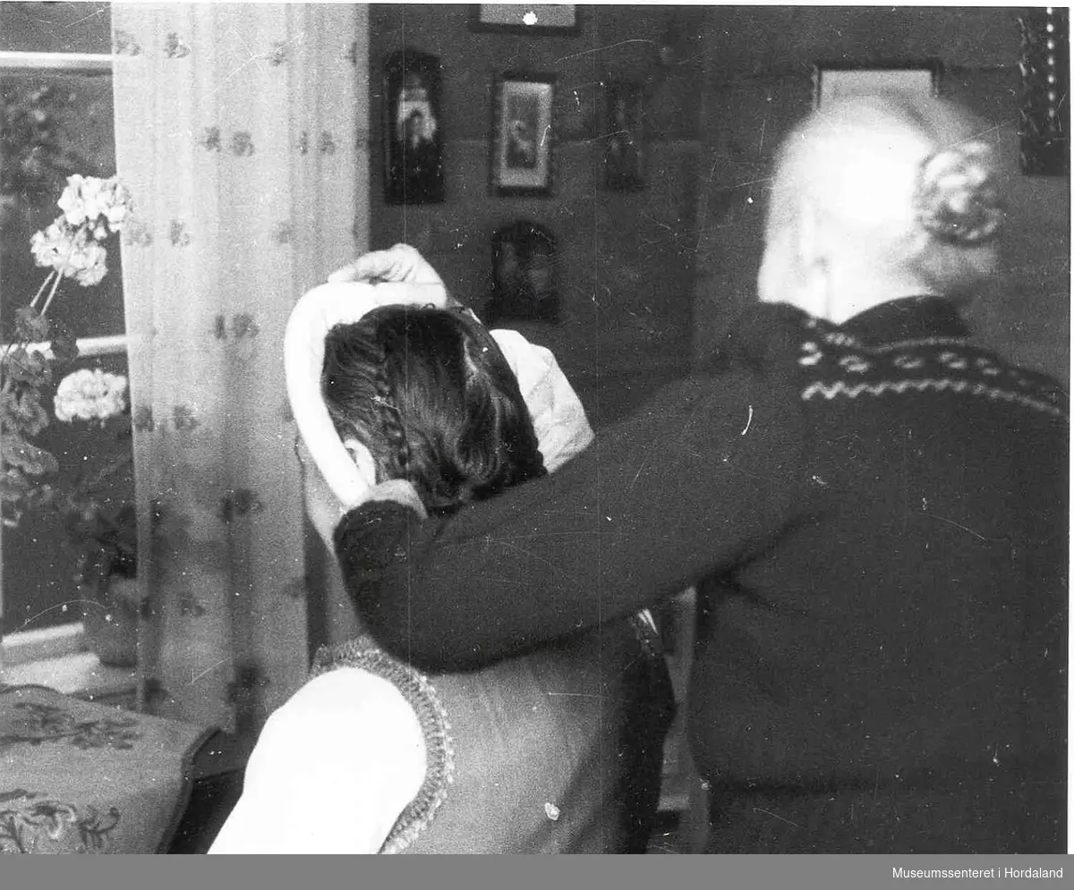 Havrå.Ei elder kone hjelper ei bunadskledd kvinne på med linhua. Her vert valken som er under hua sett på. Eldre stove med tømmerveggar. Fleire bilete med rame heng på veggen. Vindauge med gardin, og Granium i blomsterpotte i karmen. lite bord med duk på under vindauga.