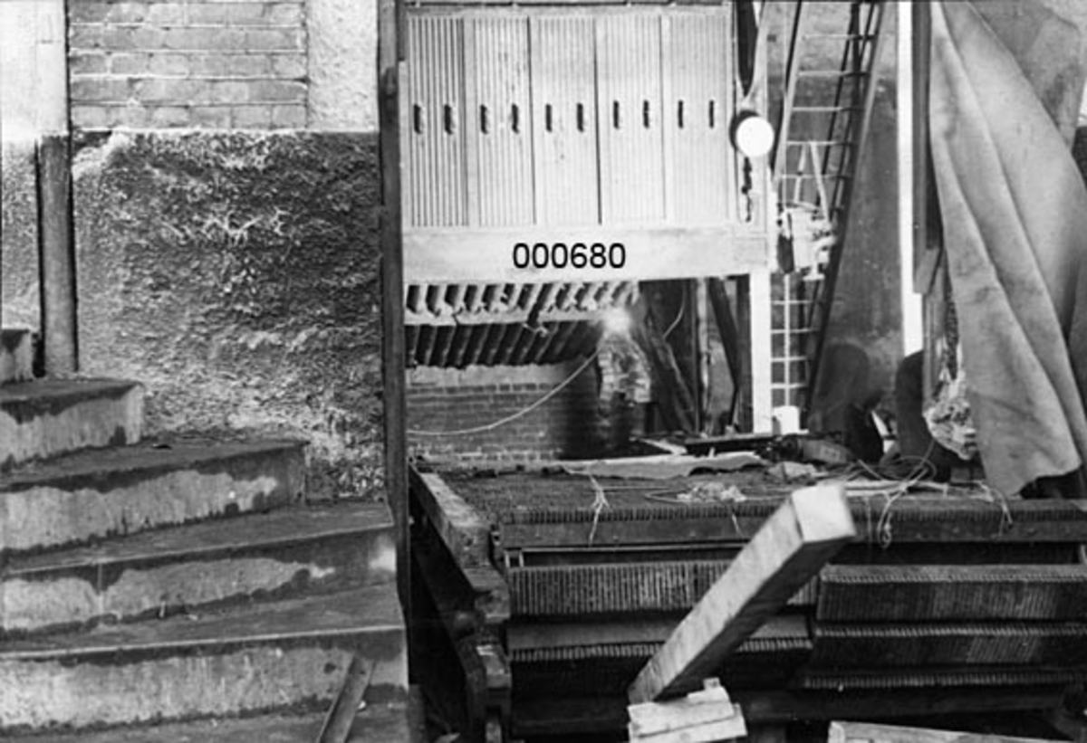 Interiør fra A/S Buskerud Papirfabrikk på Åssida i Drammen (den gang i Lier kommune).  I 1937 ble papirmaskinen ved denne fabrikken fullstendig ombygd, særlig for å kunne kjøre maskinen med større driftshastigheter og dermed oppnå større produksjonsvolumer.  Fotografiet er tatt under ombyggingsarbeidet, mot stokeren i det delvis nedrevne fyrhuset.  I den perioden fabrikken ble drevet med dampkraft ble brensel (kol) ført inn i fyrrommet ved hjelp av de avbildete ristene.  Over ristene ser vi fronten på dampkjelen. Til høyre ser vi hvordan nedrivingsveggen har vært skjermet med en presenning.  Til venstre ei støpt trapp. 